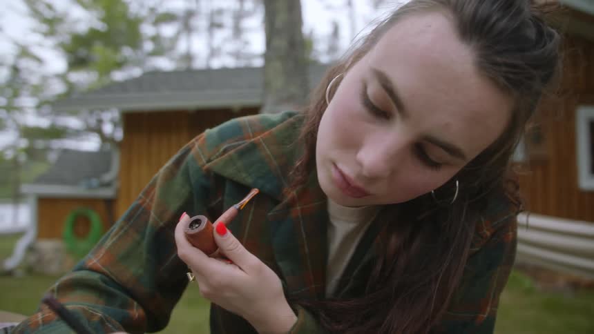 Female artist holds smoke pipe and draws while sitting at table in house yard near water