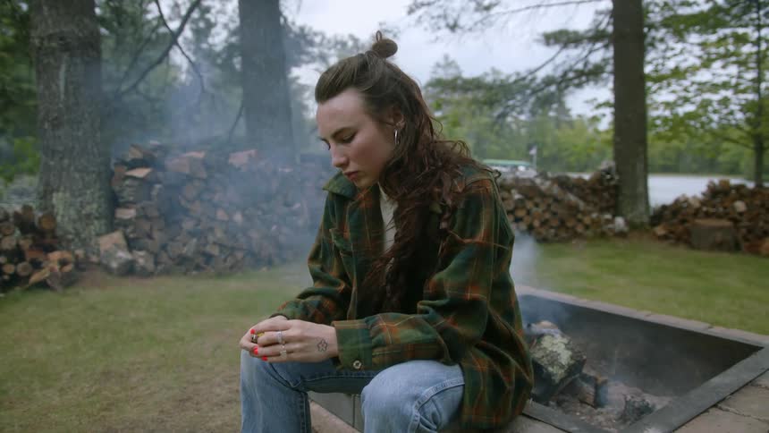 Woman sitting on the edge of fire pit in the yard near cabin in the woods by the lake