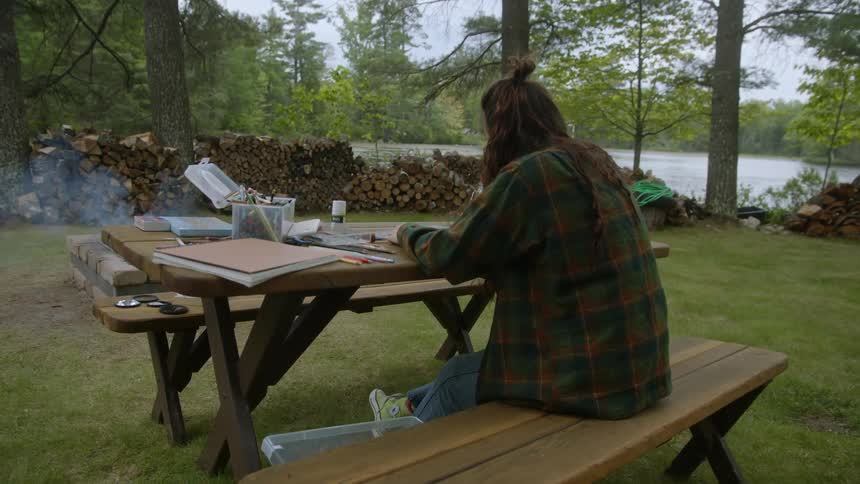 A woman draws while sitting at a picnic table along a lake