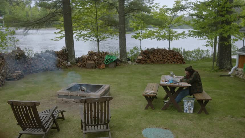 A woman outside in yard at lake housecabin in the woods sitting at table drawing while cooking something on the grill