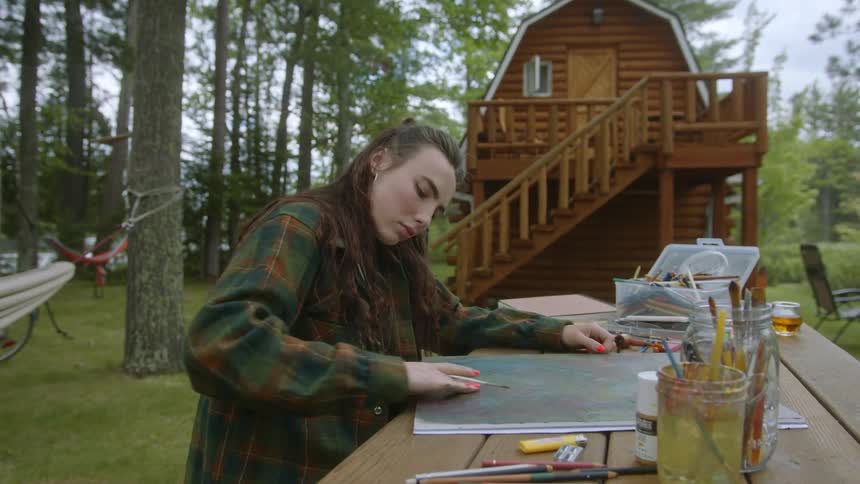 Young woman sitting on a table outside her small cabin in the woods painting with her fingers