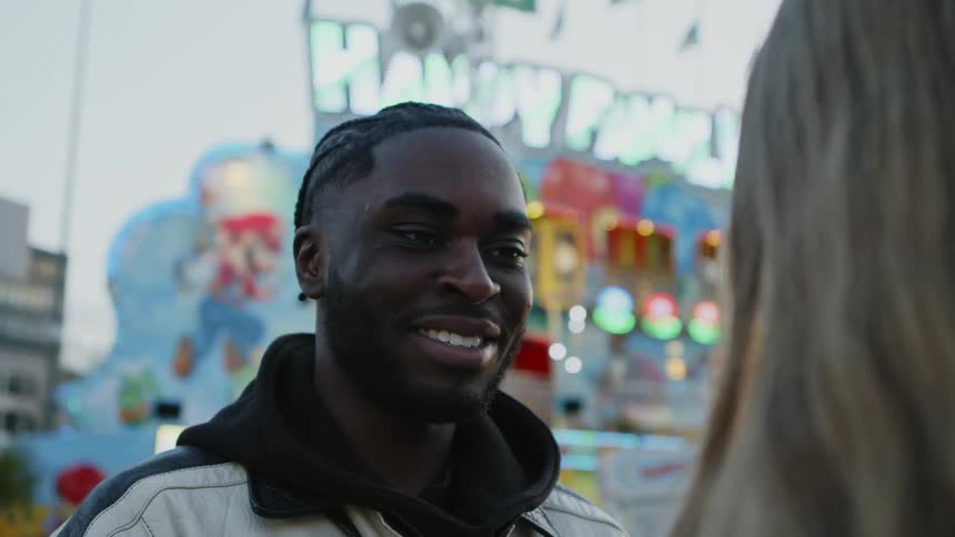 A young man talks to a woman at a fair with a mobile carnival game with flashing lights in the background