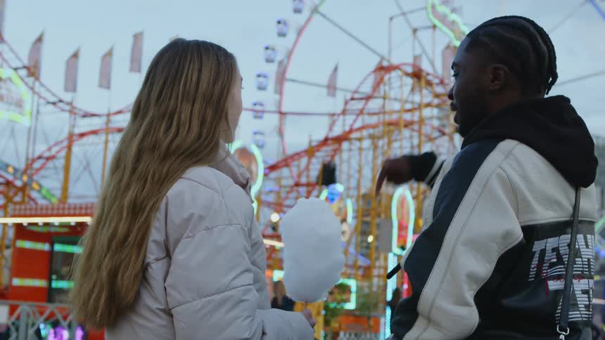 Young woman and man on a date at a carnival early evening he is talking to her and pointing at things