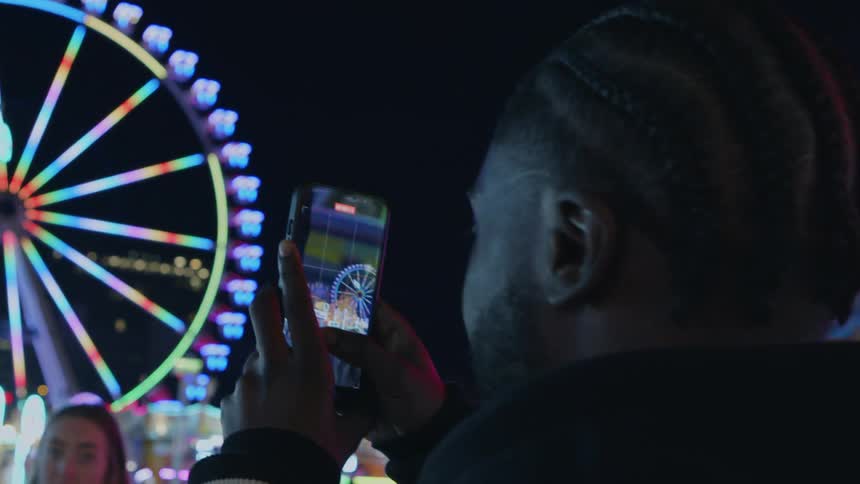 Man taking video of ferris wheel at nighttime while female date watches in the background