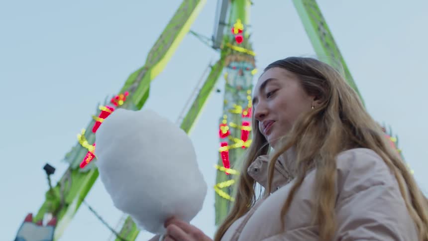 A woman eats cotton candy while standing near a carnival ride