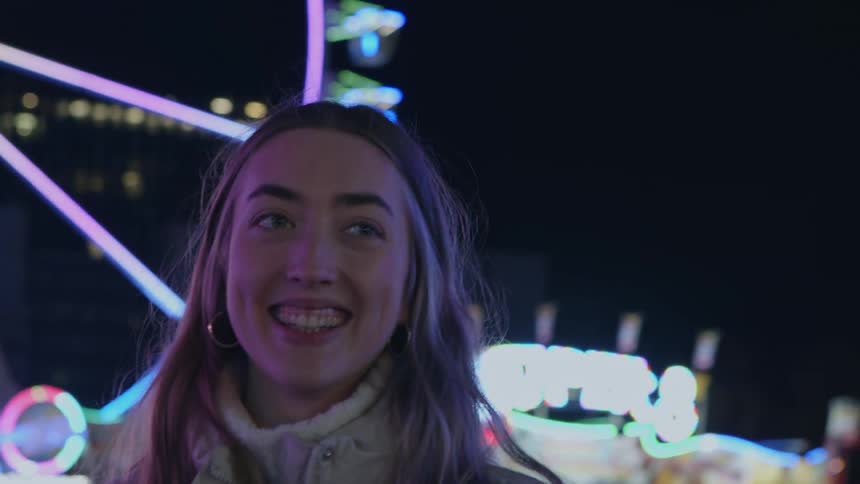 Blonde woman with earrings and beige coat smiles at the amusement park