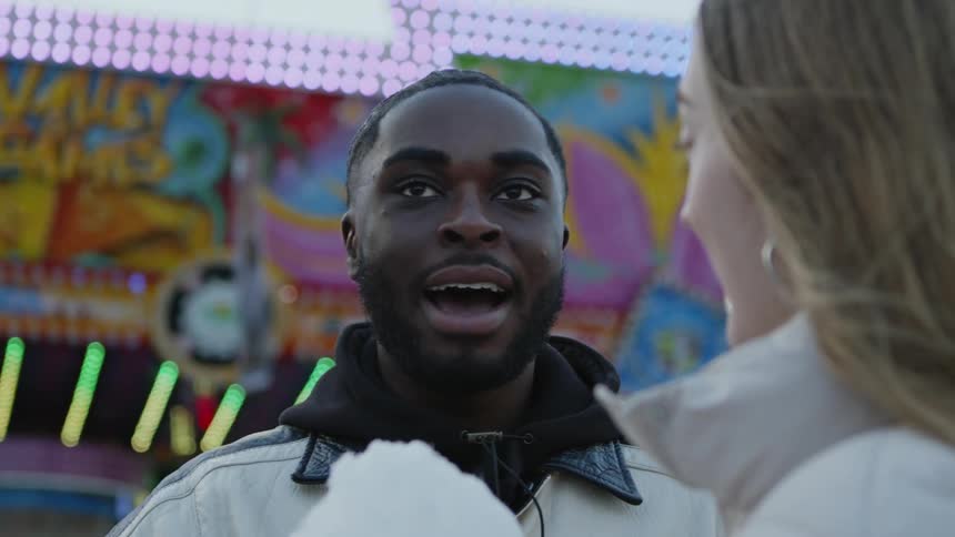 A man talking to his date in front of him while she hold cotton candy and they stand near fair games