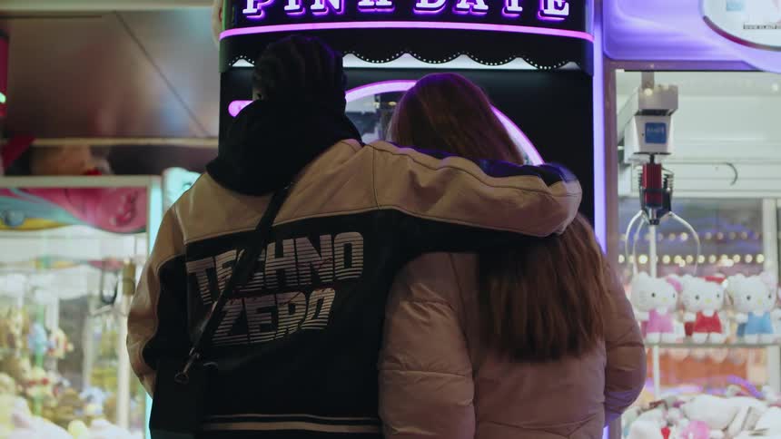 The backside of a man and woman standing in front of an arcade game the man with his arm around the woman