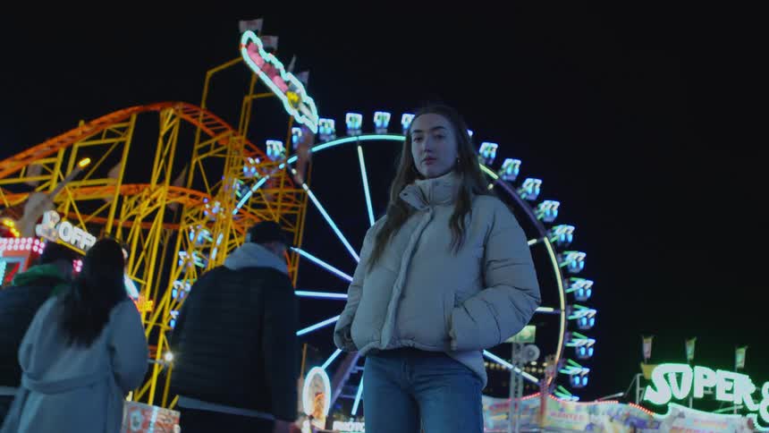 Woman posing for photo in front of ferris wheel at nighttime carnival people passing by