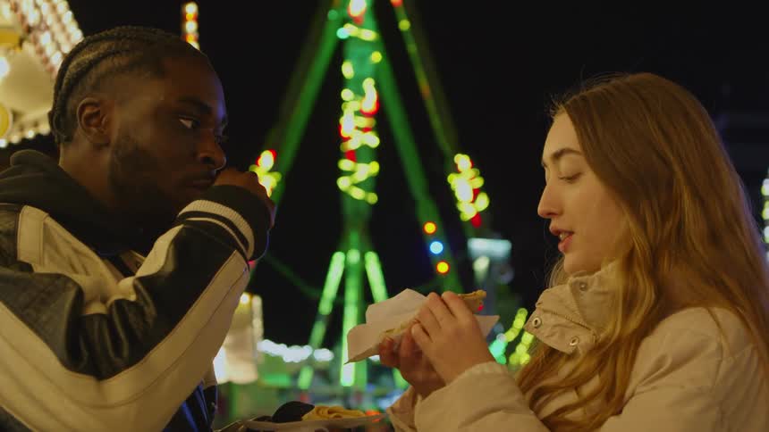 Man and woman stand eating and talking at amusement park at night