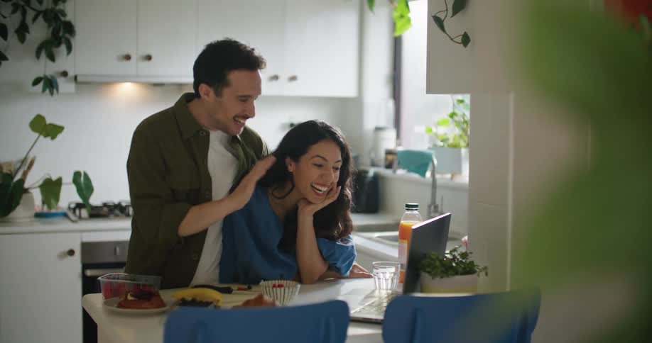 A man and woman at their kitchen table laugh as they talk to someone through a laptop