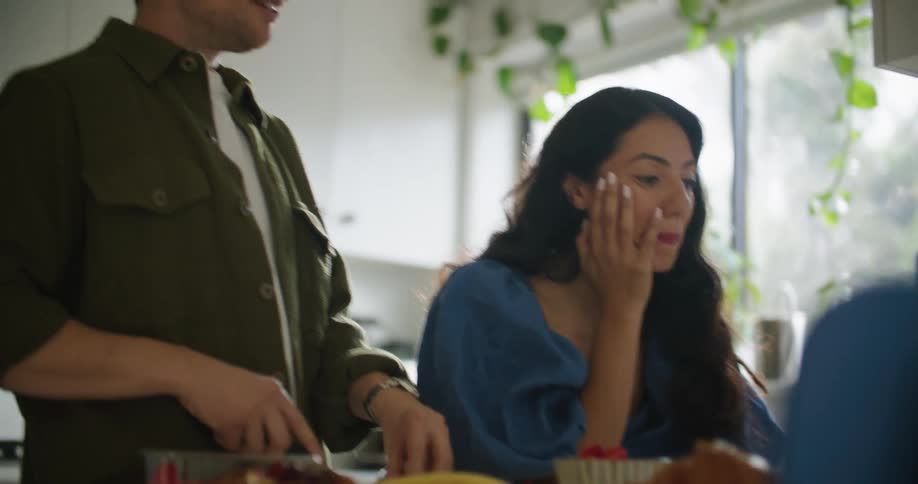 Man with mustache and green shirt cuts fruits while woman with wavy hair and bleu shirt make video call on laptop in the kitchen