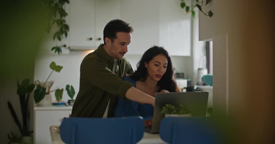 A couple looks at a laptop screen together while standing in the kitchen