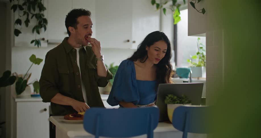 A man and woman standing in their kitchen eating and looking at a laptop