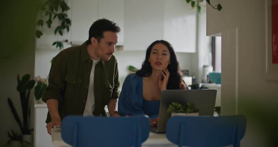 A man and woman in the kitchen looking at a laptop together and discussing something