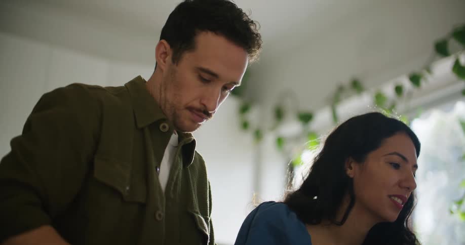 Woman with dark wavy hair blue shirt and man with green shirt and mustache look at laptop in the kitchen