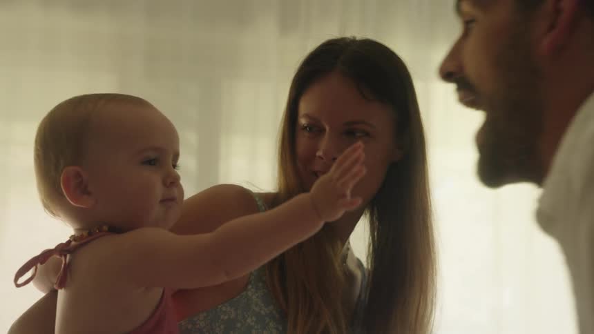 A mother holding her baby daughter as the father makes funny faces at the baby and smiling