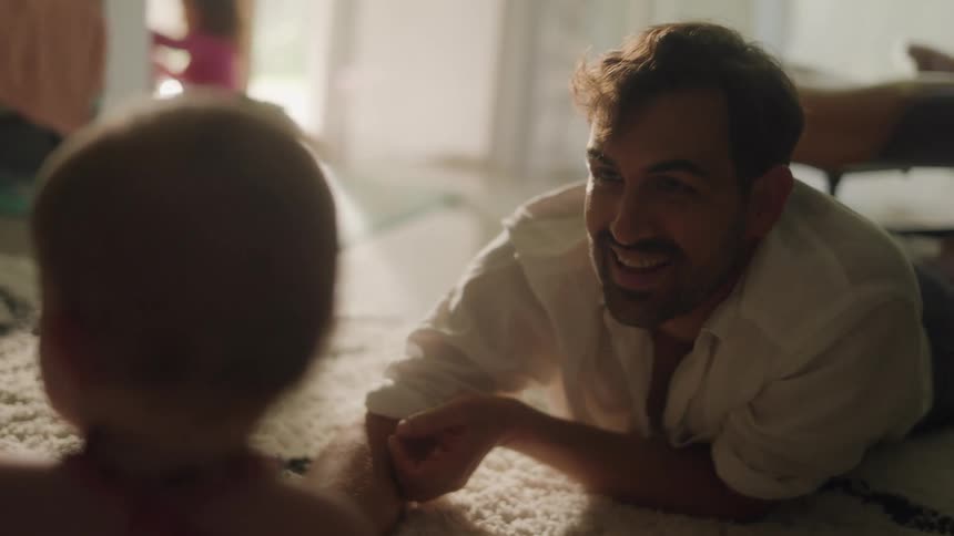 A father lays on the floor playing and smiling with his baby child with a little girl playing in the background