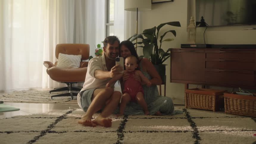 A mother and father sit on the floor holding their baby and holding out their smartphone in front of them for a photo or video