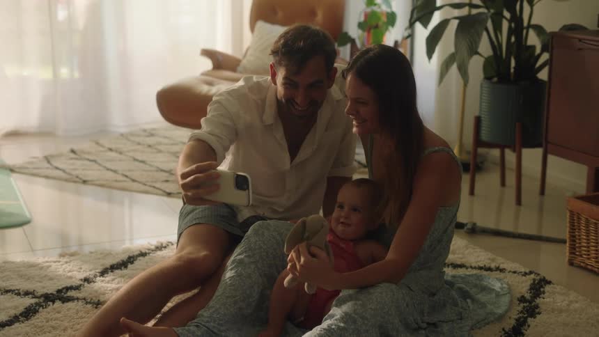 Mom dad and baby girl pose together on the floor at home for a selfie