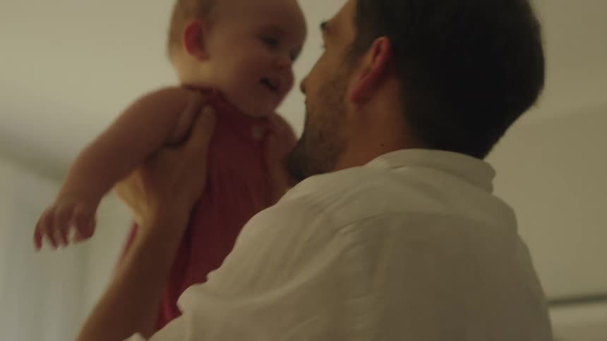 Father with stubble beard and white shirt holds up toddler girl in the living room
