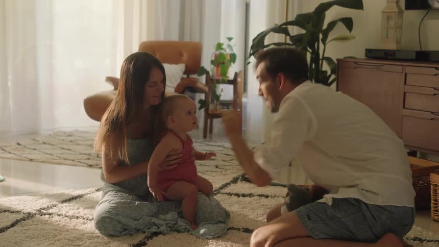 Father with stubble beard white shirt and shorts play with toddler daughter sitting on mothers lap in the living room at home