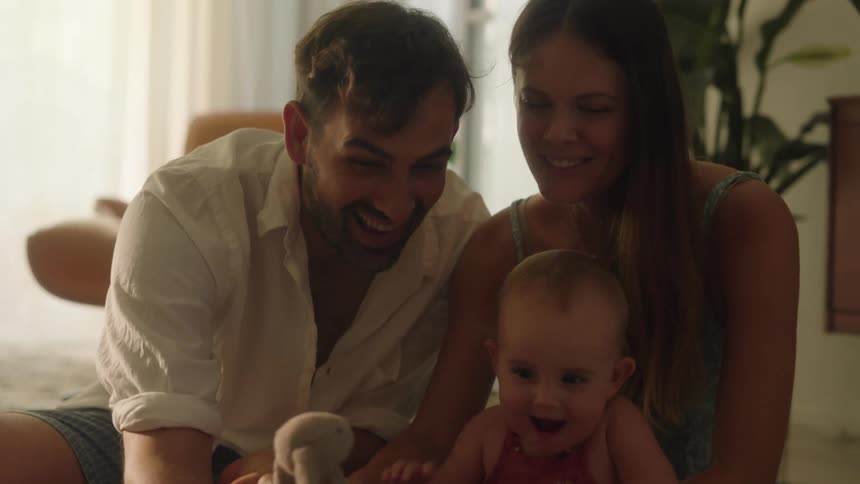 Father with white shirt shorts and mother with straight hair and green dress and toddler daughter take a selfie with smartphone in the living room at home