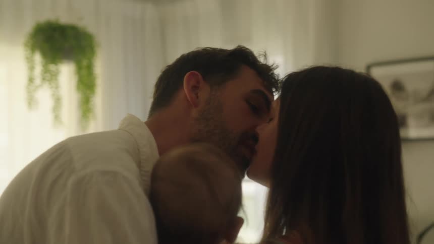 Mom and dad kissing romantically while holding their baby at home during the day