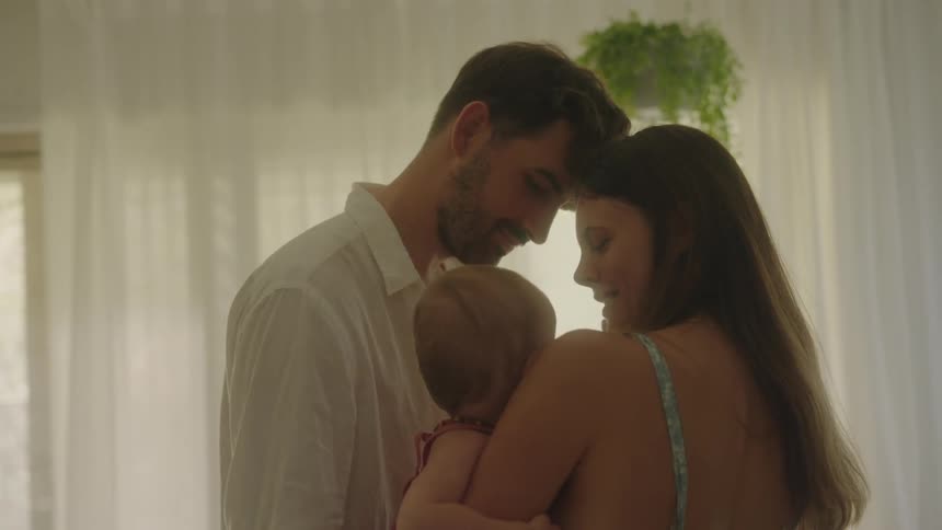 A mother holding her baby as the father stands there kissing the babies head in a bright room