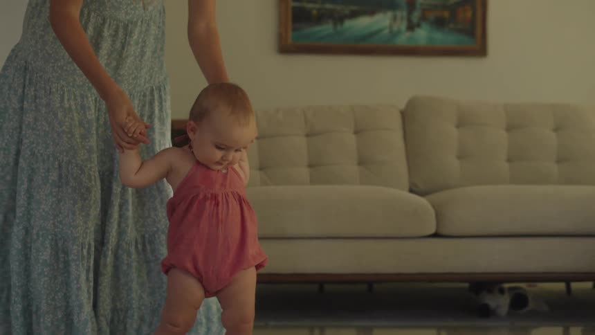 A mother walks her baby daughter in their living room by holding her hands
