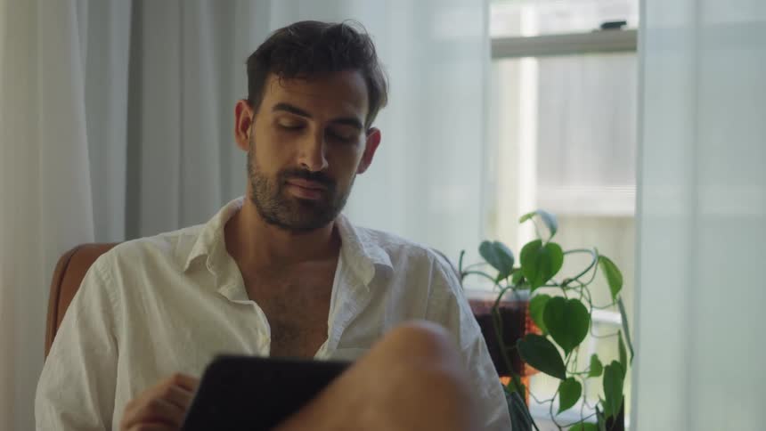 Young man sits comfortably at home next to a window browsing a tablet