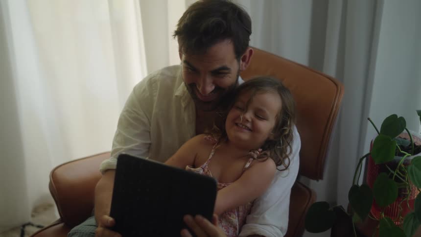 Daddy and daughter sitting together at home looking at a tablet laughing and smiling