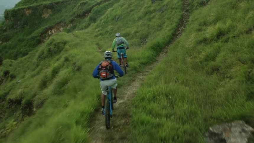 A pair of cyclists ride along a narrow path on a mountainside during the day