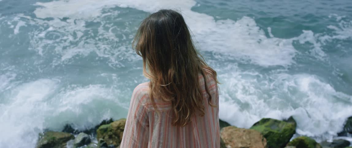 Woman standing at edge of cliff overlooking waves crashing on rocky shore