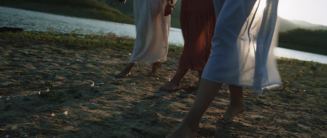 Group of women walking on the sand near a river with their dresses waving in the wind at morning
