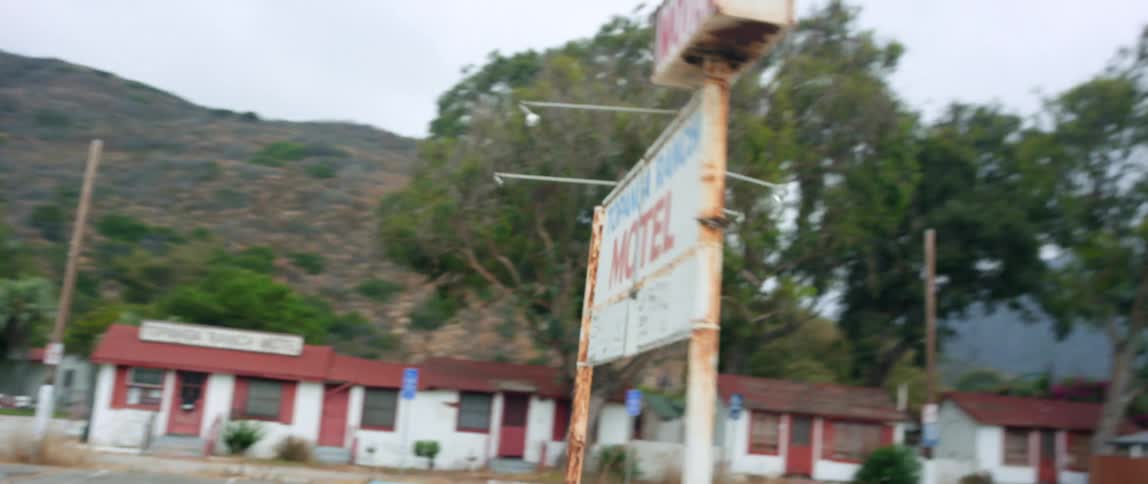 During a road trip vehicle passes nearly empty parking lot near motel with mountains in the background