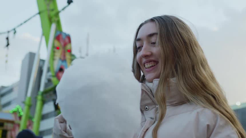 A young girl eating cotton candy outdoors with a fair behind her