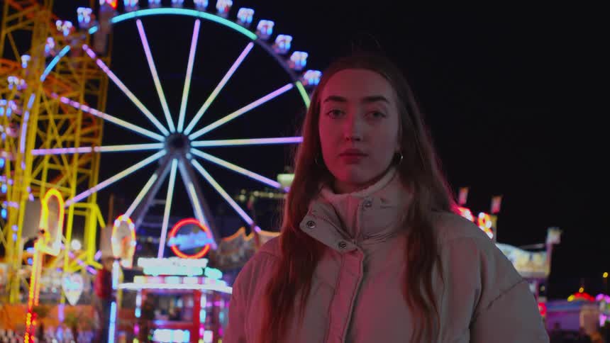 Young woman wearing a thick jacket standing in front of an amusement park at night
