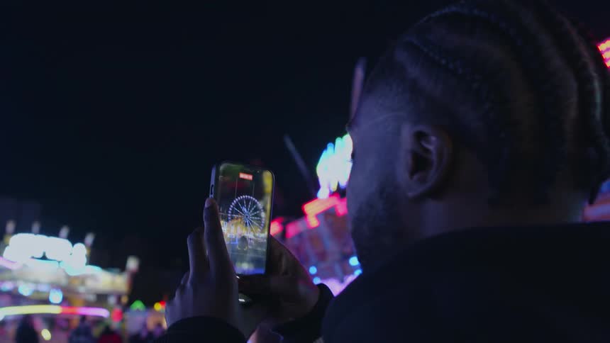 A man holds up his phone to capture images of a brightly illuminated ferris wheel