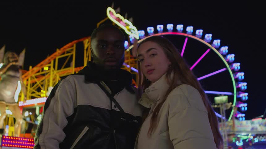 Young couple in front of a ferris wheel on an amusement park looking at the camera