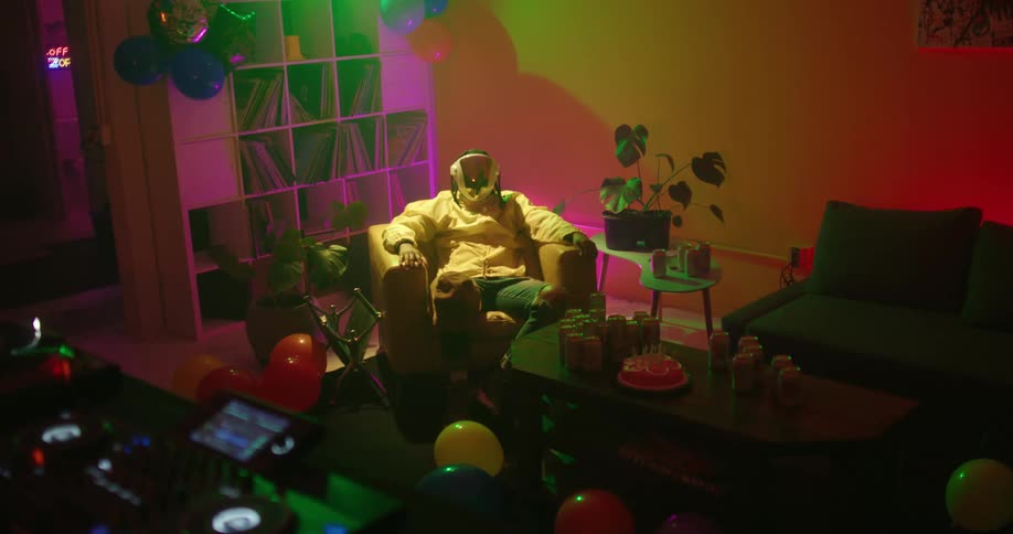 A person sitting in a chair wearing a yellow suit with balloons on the floor and cans on the table in front of him