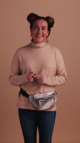 A woman wearing a fanny pack over her sweater and jeans stands laughing and smiling and looking around