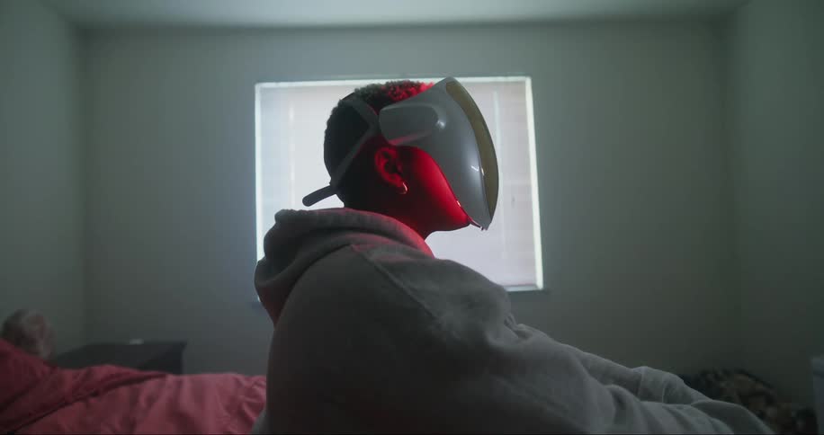 A man wears a red light therapy mask while sitting in bed