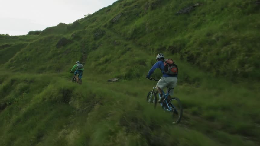Two men on bicycles ride on along a cliff ledge on a grassy mountain