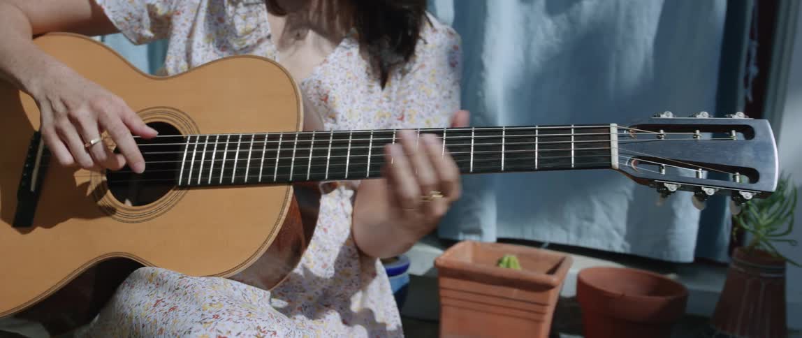 A woman wearing a dress is moving her hand up and down the neck of an acoustic guitar as she plays