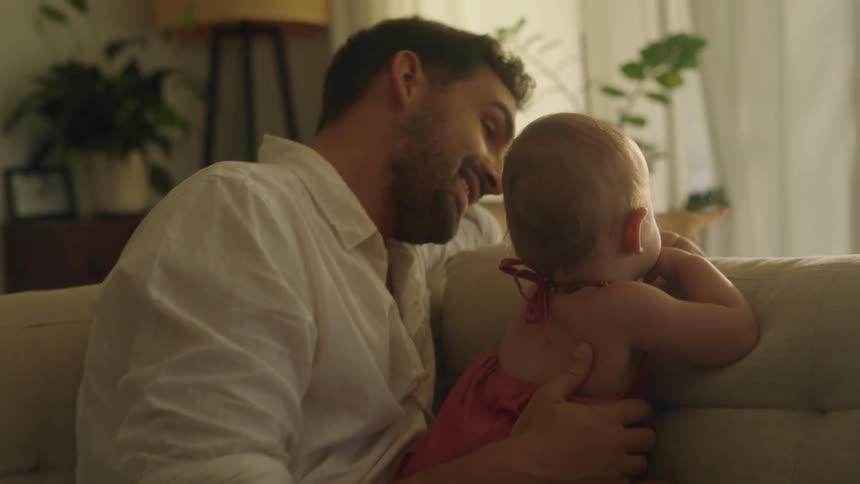 A man is sitting on the living room couch holding his baby and pointing to the window as he speaks