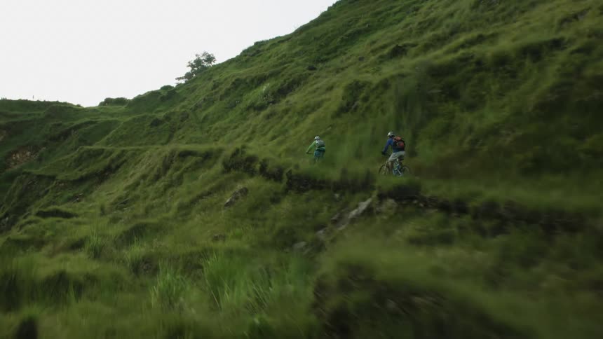 A pair of cyclists ride along a grassy mountainside at high speed