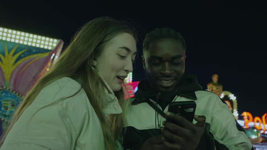Young couple looking at pictures in a smartphone standing in the middle of an amusement park at night
