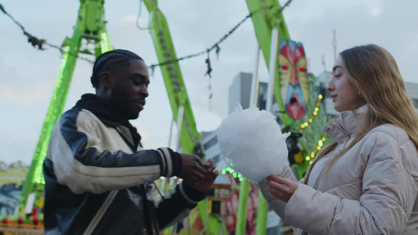 A young man and woman share some cotton candy at an amusement park