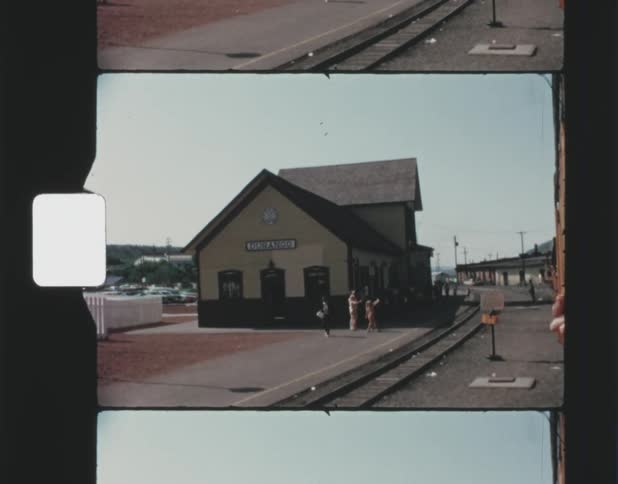 Pedestrians walk along area around train terminal between train tracks and parking lot with hills in the distance